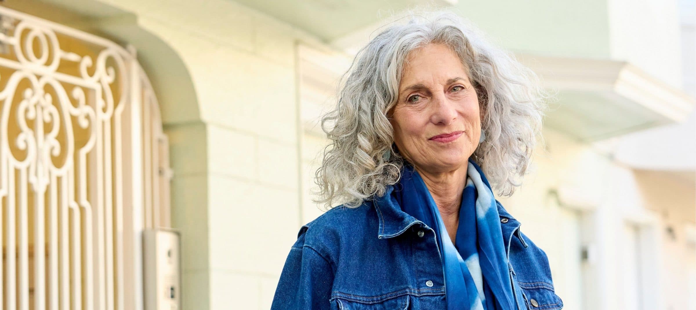 Smiling older woman in a denim jacket standing outside her home